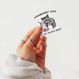 May include: A white sticker with a black and white illustration of a raccoon holding a glass of strawberry jam. The text on the sticker reads "Strawberry Jams But My Glock Don't".