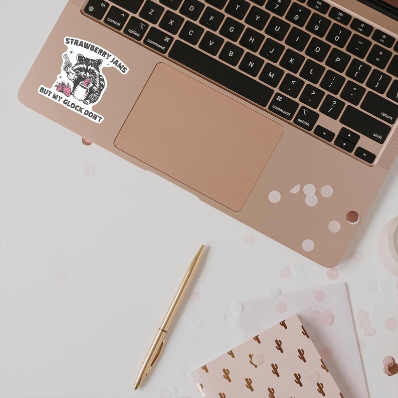 May include: A sticker with a raccoon holding a glass of strawberry jam. The sticker has the text "Strawberry Jams But My Glock Don't" on it. The sticker is on a laptop computer.