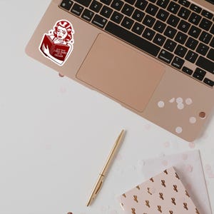 May include: A sticker of a woman with red hair reading a book with the text "Not This I Fell Hard For Villain". The sticker is on a laptop computer with a rose gold finish.