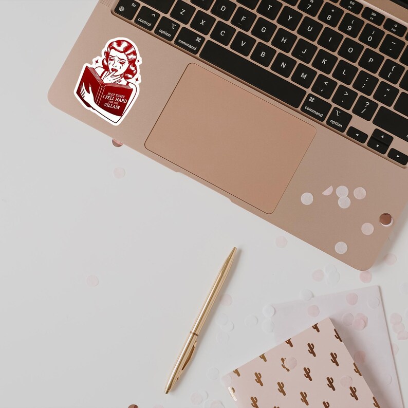 May include: A sticker of a woman with red hair reading a book with the text "Not This I Fell Hard For Villain". The sticker is on a laptop computer with a rose gold finish.