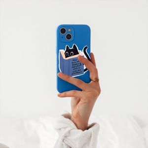 May include: A blue phone case with a black cat peeking out of a book. The book has the text "How we buy new books & pretend like it was an accident".