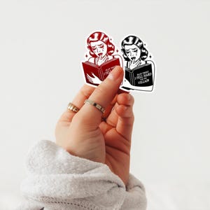 May include: Two stickers featuring a woman with tears in her eyes reading a book. The text on the book reads "Plot Twist I Fell Hard For The Villain". One sticker is red and the other is black and white.