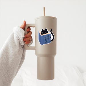 May include: A beige travel mug with a black cat sticker on it. The sticker shows a black cat peeking out of a blue book with the text "How to buy new books & pretend like it was an accident".