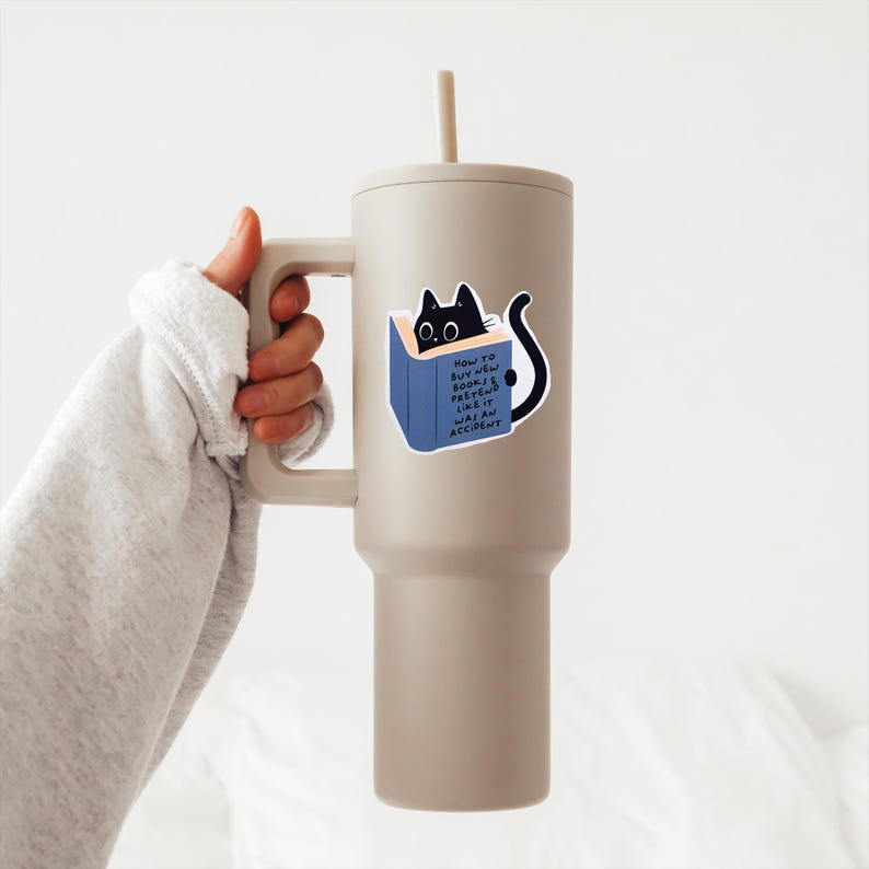 May include: A beige travel mug with a black cat sticker on it. The sticker shows a black cat peeking out of a blue book with the text "How to buy new books & pretend like it was an accident".