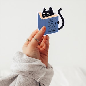 May include: A blue book sticker with a black cat peeking out from the top. The text on the sticker reads "How to buy new books & pretend like it was an accident."