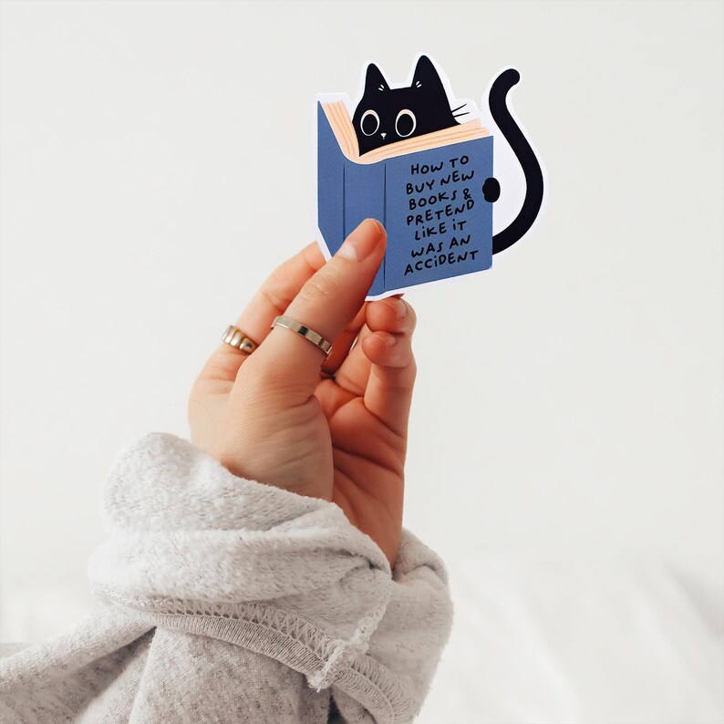 May include: A blue book sticker with a black cat peeking out from the top. The text on the sticker reads "How to buy new books & pretend like it was an accident."