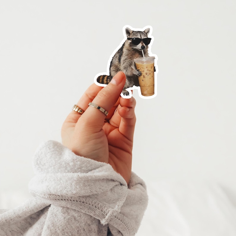 May include: A sticker featuring a raccoon wearing sunglasses and drinking iced coffee. The raccoon is gray and brown with a striped tail. The iced coffee is in a clear plastic cup with a straw. The sticker is held up by a hand.