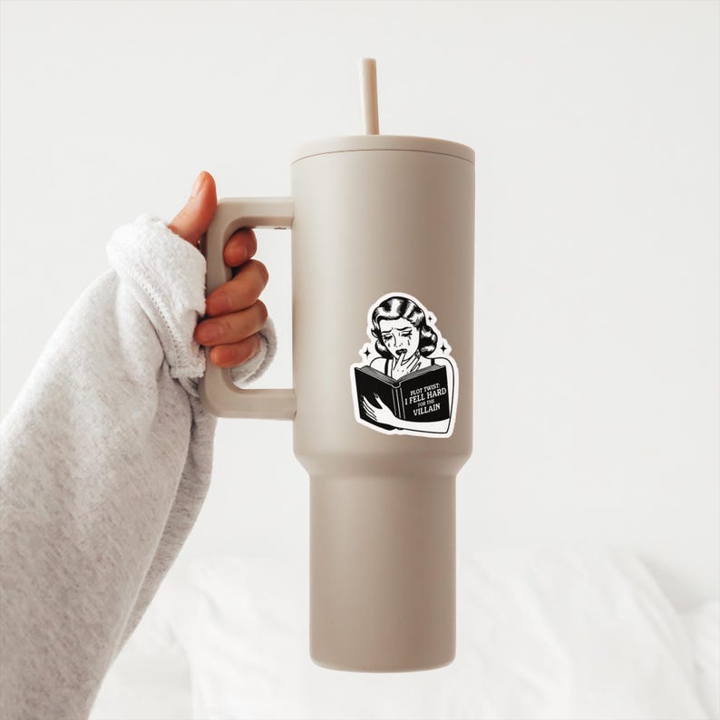 May include: A beige stainless steel tumbler with a black and white sticker featuring a woman reading a book with the text "Must Trust I Feel Hard For The Villain".