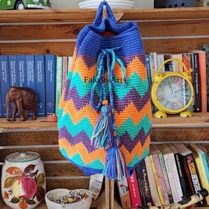 Puede incluir: Una mochila de ganchillo con cierre de cordón que presenta un vibrante patrón de chevrón en rayas horizontales de color azul, naranja, morado y turquesa. La bolsa incluye un cordón trenzado azul con dos borlas decorativas y el texto Falcon Arts.