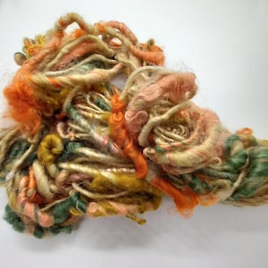 Könnte beinhalten: Ein Knäuel handgesponnener Wolle in Orange-, Grün- und Beigetönen. Die Wolle besteht aus Naturfasern und hat einen rustikalen, handgefertigten Look.