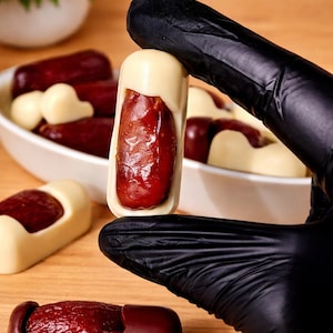 May include: A close-up of a date fruit encased in white chocolate, held by a gloved hand. Other chocolate-covered dates are in a white bowl and on a wooden surface. The dates have a rich, dark red color.