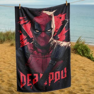 Może przedstawiać: Czarny ręcznik plażowy z czerwono-czarną grafiką Deadpoola. Projekt zawiera twarz Deadpoola, dwa miecze i napis "DEAD POOL" w kolorze czerwonym. Ręcznik wisi na sznurku na piaszczystej plaży.
