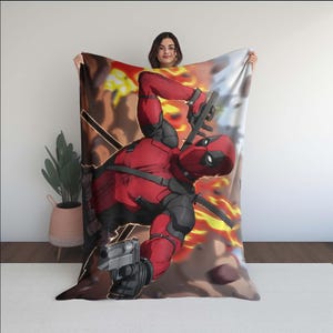 Puede incluir: Un personaje de Deadpool rojo y negro con una pistola en la mano, impreso en una manta de forro polar. La manta tiene un fondo amarillo y naranja con llamas.