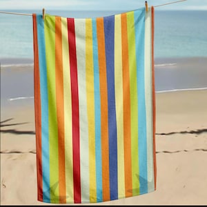 Op de afbeelding: Een gestreepte strandlaken met verticale strepen in de kleuren rood, oranje, geel, groen, blauw en wit. De handdoek hangt aan een waslijn, met een strand en de zee op de achtergrond. De handdoek is gemaakt van zacht, absorberend materiaal.