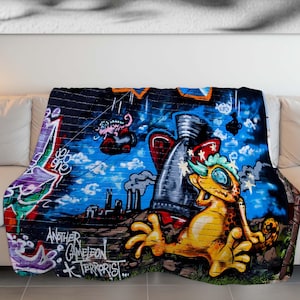 May include: A colorful throw blanket with a graffiti design featuring a yellow lizard, a rocket, and the text "Another Chameleon Terrorist".