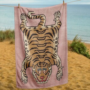 May include: A pink beach towel with a brown and black tiger printed on it. The tiger is lying on its back with its paws in the air. The towel is hanging on a clothesline.