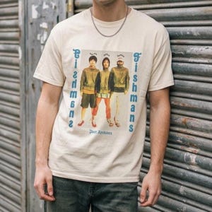 以下が含まれることがあります： クリーム色の半袖Tシャツで、ジャケットを着た3人の人物のグラフィックプリントが施されています。画像の両サイドには「Fishmans」という文字が青色で縦にプリントされており、人物の下には「Poet Archives」という文字が青色で記されています。