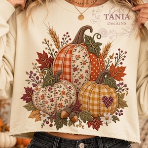 Puede incluir: Una sudadera de manga larga color crema con un gráfico de tres calabazas con varios patrones, incluyendo cuadros, flores y lunares, rodeadas de hojas de otoño, trigo y bellotas. El texto Bella TANIA DesiGNS está impreso en la parte superior.