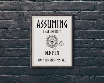 May include: A framed wall print with a white background and black text that reads: ASSUMING I WAS LIKE MOST OLD MEN WAS YOUR FIRST MISTAKE. The design includes a circular Viking-style shield and a battle axe illustration centred between the text lines.