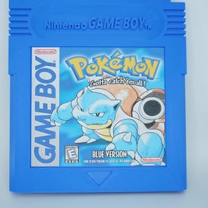May include: Blue Nintendo Game Boy cartridge for the game Pokémon Blue Version. The cartridge features the Pokémon logo and a graphic of a blue turtle-like creature. The text "Gotta catch 'em all!" is also visible.