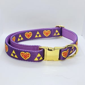 May include: A purple dog collar with a gold-coloured buckle and D-ring. The collar has a repeating pattern of yellow triangles and red heart-shaped designs. The collar is made of a durable fabric.