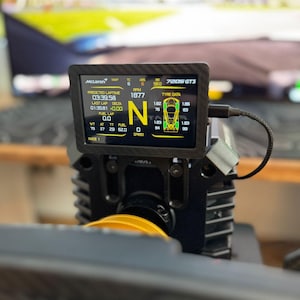 以下が含まれることがあります： 「McLaren」と「720S GT3」の文字が表示された、ラップタイムやRPMなどのレースデータを示す黒いデジタルディスプレイ。画面は、黄色のホイールが付いた黒いデバイスに取り付けられています。測定値はメートル法です。