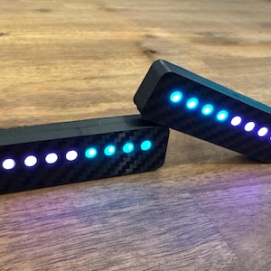 Könnte beinhalten: Zwei schwarze, rechteckige Geräte mit Karbonfasertextur, die jeweils eine Reihe von blau und weiß leuchtenden Lichtern anzeigen. Die Geräte befinden sich auf einer Holzoberfläche, wobei die Lichter in einem Muster leuchten.