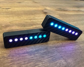 May include: Two black rectangular devices with a carbon fiber texture, each displaying a series of glowing blue and white lights. The devices are positioned on a wooden surface, with the lights illuminating in a pattern.