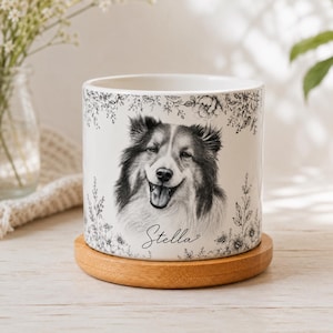 May include: A white ceramic cylindrical planter featuring a black and white portrait of a dog with a smiling expression. The design includes delicate black floral illustrations surrounding the dog and the name Stella written in cursive script.