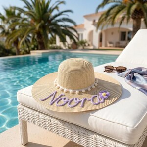 Peut inclure: Un chapeau de soleil en paille beige avec une bordure à pompons blancs autour de la base de la calotte. Le bord est orné du prénom Noora en lettres de laine violette et d'une petite fleur crochetée violette au centre jaune sur le côté.