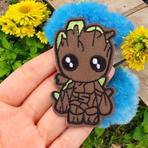 Peut inclure: Un écusson brodé marron représentant un personnage de dessin animé avec de grands yeux et des détails verts. L'écusson est tenu dans une main, avec un accessoire bleu duveteux en arrière-plan et des fleurs jaunes.