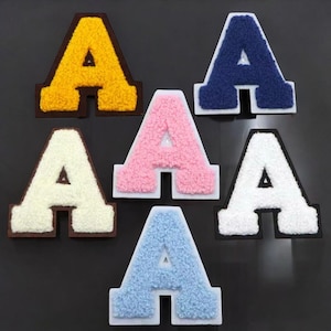 Puede incluir: Un conjunto de seis parches con la letra "A" en varios colores. Los parches tienen una textura difusa y son de color amarillo, azul marino, rosa, blanco y azul claro. Cada letra está delineada en un color contrastante.