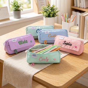 May include: Five rectangular pencil cases in pastel colors including mint, lavender, light blue, and pink. Each case features a front flap with a small circular button, a zipper closure, and a white label with the text HONG TWAN. Names are embroidered on the flaps.