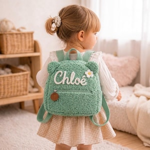 Peut inclure: Un sac à dos en sherpa vert sauge avec des oreilles d'ours, une marguerite brodée blanche et le prénom Chloe en lettres cursives blanches. Le sac comprend une poche avant zippée avec une tirette ronde marron et des bretelles assorties.