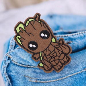 Puede incluir: Un parche bordado marrón con un personaje de dibujos animados con apariencia de árbol. El parche tiene un cuerpo marrón, detalles verdes y ojos negros y blancos. El parche está colocado sobre una superficie de mezclilla azul.