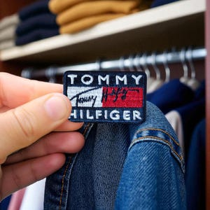 Op de afbeelding: Een rechthoekige patch met het Tommy Hilfiger-logo in wit, rood en blauw. De patch wordt voor een spijkerjasje gehouden. Het logo bevat de merknaam en een gestileerd vlagontwerp.
