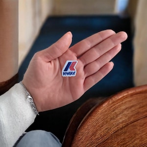 Può includere: Una piccola toppa quadrata con la scritta "KWAY" in blu e un design a freccia rosso e blu. La toppa è bianca con un bordo blu. La toppa è tenuta in una mano aperta.