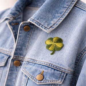 Puede incluir: Primer plano de una chaqueta vaquera azul claro con un parche de trébol de cuatro hojas. El parche es verde y amarillo, y la chaqueta tiene botones y bolsillos dorados. Se ve el cuello de la chaqueta.
