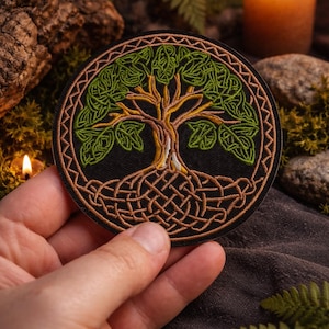 Può includere: Toppa rotonda ricamata con un disegno dell'albero della vita celtico. La toppa ha uno sfondo nero con un albero dettagliato in tonalità verdi e marroni. Le radici e i rami dell'albero sono intrecciati in un motivo a nodo.