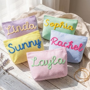 Könnte beinhalten: Fünf rechteckige Reißverschlusstaschen aus Seersucker-Stoff in Pastellfarben wie Lila, Gelb, Grün, Blau und Rosa. Jede Tasche ist mit einem Namen in einer Kontrastfarbe bestickt: Linda, Sunny, Sophia, Rachel und Layla.