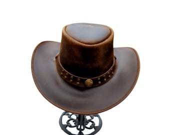 Puede incluir: Un sombrero vaquero de cuero marrón con un ala ancha y una banda decorativa con tachuelas metálicas. El sombrero se muestra en un soporte de metal negro.