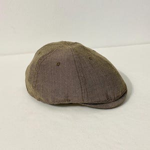 May include: A brown herringbone pattern flat cap. The cap has a rounded crown and a small, stiff brim. The fabric appears to be a textured, woven material. The cap is a neutral color, suitable for various outfits.