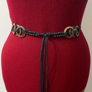 May include: A brown and black belt with a macrame design. The belt features two decorative, circular clasps with a woven pattern. The belt is tied with a knot and has long, dangling black cords. The belt is displayed on a red garment.