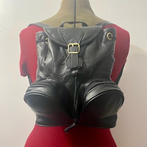 May include: Black leather backpack with a flap closure and a gold-colored buckle. The backpack has two front zippered pockets and quilted stitching details. The bag is displayed on a mannequin with a red top.