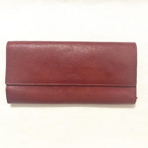 May include: A burgundy leather wallet with a textured surface. The wallet has a rectangular shape with a fold-over flap. The wallet is closed and is set against a white background.