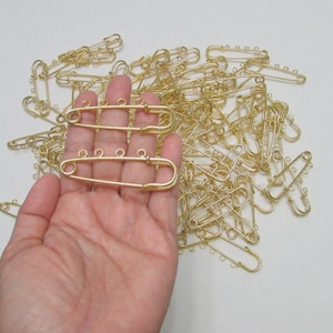 May include: A collection of gold-colored safety pins, some held in a hand. The pins have a unique design with small loops for attaching charms or beads. These pins are ideal for jewelry making or craft projects.