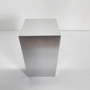 Pode incluir: Um bloco de metal prateado, de forma retangular, com um acabamento escovado. O bloco está em posição vertical sobre uma superfície branca. A superfície superior é plana e lisa. As bordas do bloco são afiadas e bem definidas.