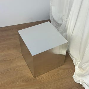 Peut inclure: Une table d'appoint carrée dotée d'une finition en métal argenté poli et réfléchissant. L'objet présente des bords nets et précis ainsi qu'une surface supérieure lisse et plane, formant un cube géométrique qui reflète son environnement.