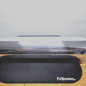 Pode incluir: Um apoio de pulso cromado brilhante e um tapete de rato preto com o nome da marca "Fellowes" impresso em branco. O apoio de pulso é cilíndrico e reflete a luz. O tapete do rato é oval.
