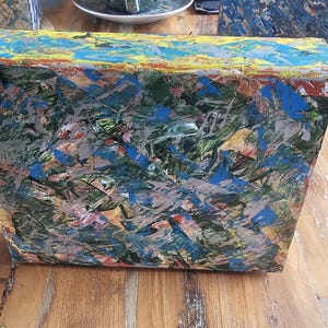 Könnte beinhalten: Eine kleine, rechteckige Holzkiste mit einem farbenfrohen abstrakten Gemälde auf der Vorderseite. Das Gemälde zeigt eine Mischung aus blauer, grüner, gelber und roter Farbe, die eine dynamische und strukturierte Oberfläche erzeugt.